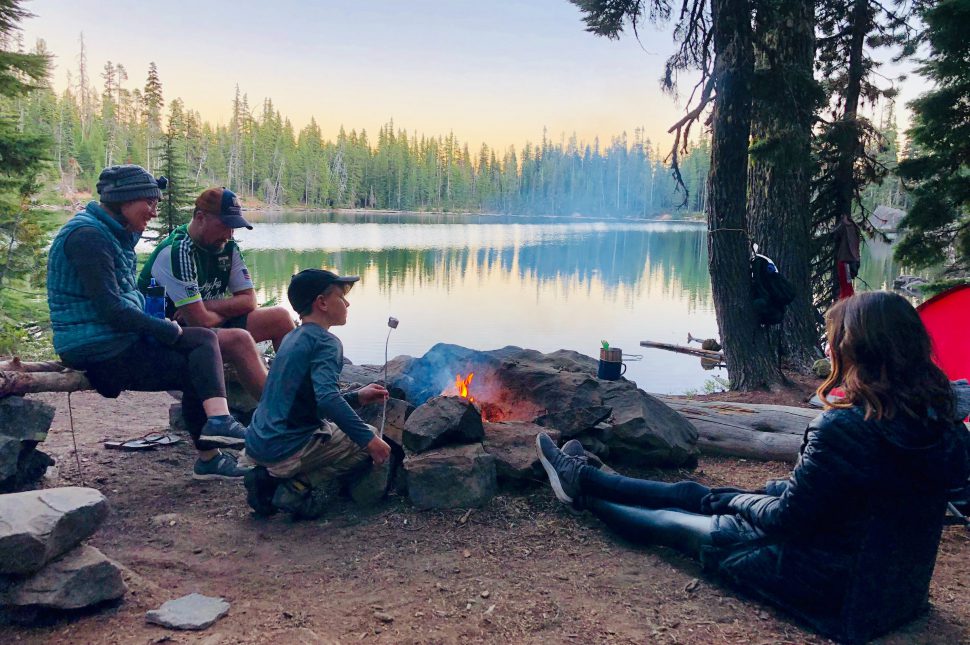 A family cooking marshmallows over a fire while camping by a lake and learning how to camp.